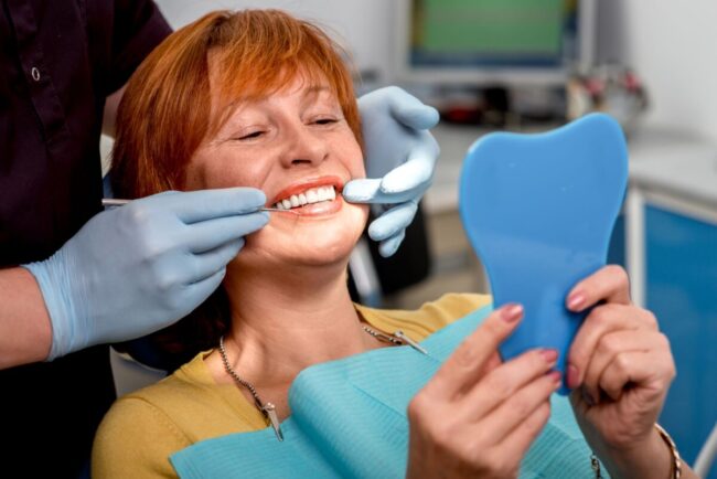 Choosing dental bridges or dentures, as represented by this woman in a dentist chair with straight white teeth, depends on your budget and your existing teeth.